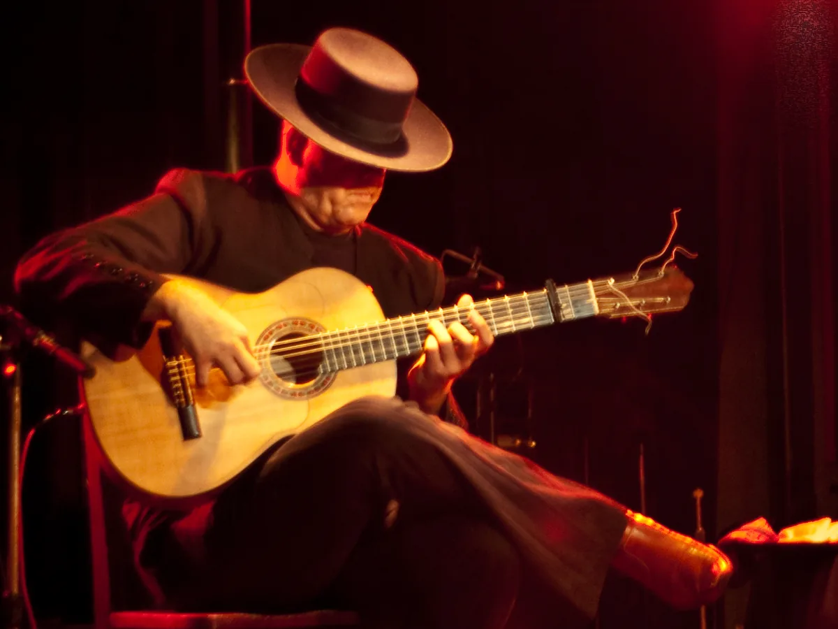 A flamenco guitarist in a wide-brimmed hat playing seated on a dimly lit stage